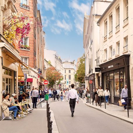 Rue animée avec des gens, cafés en terrasse et bâtiments typiques sous un ciel bleu.