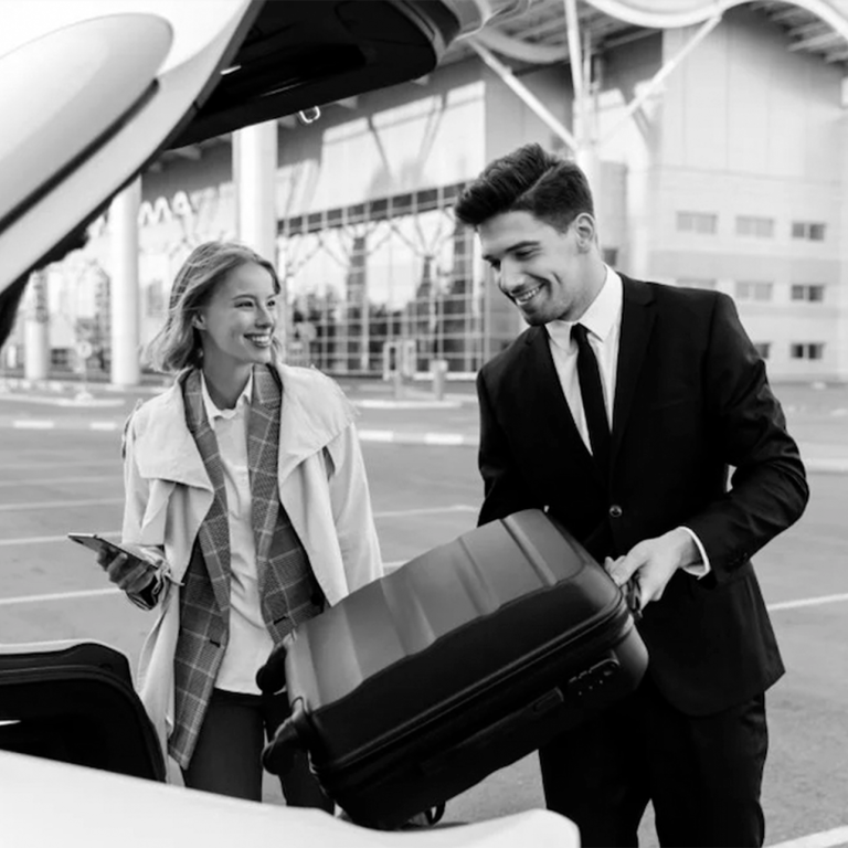 Homme en costume chargeant une valise dans le coffre d'une voiture, femme souriante à ses côtés.