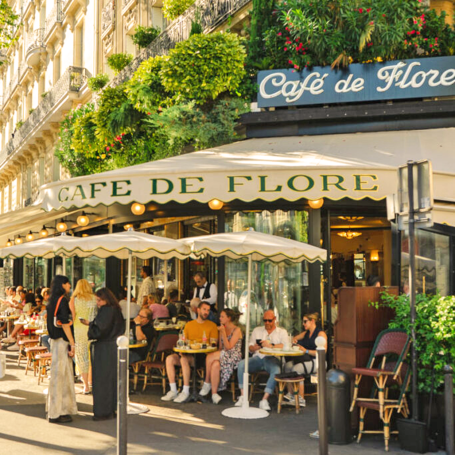 Café de Flore avec des clients en terrasse à Paris, entouré de verdure.