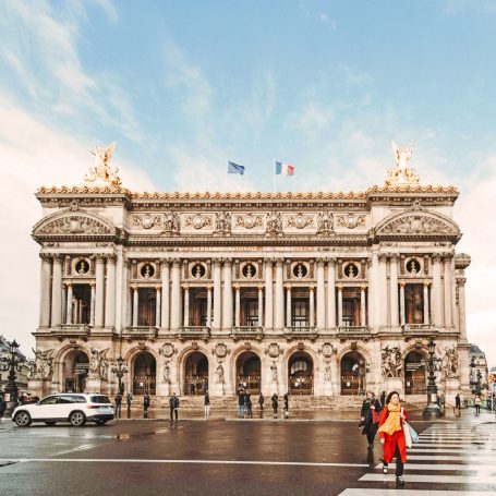 Grand bâtiment architectural avec drapeaux, devant un espace urbain animé.