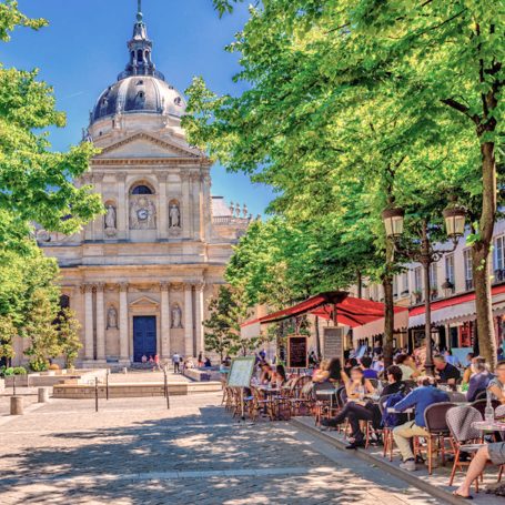 Terrasse animée devant un bâtiment historique, entourée d'arbres et de verdure.