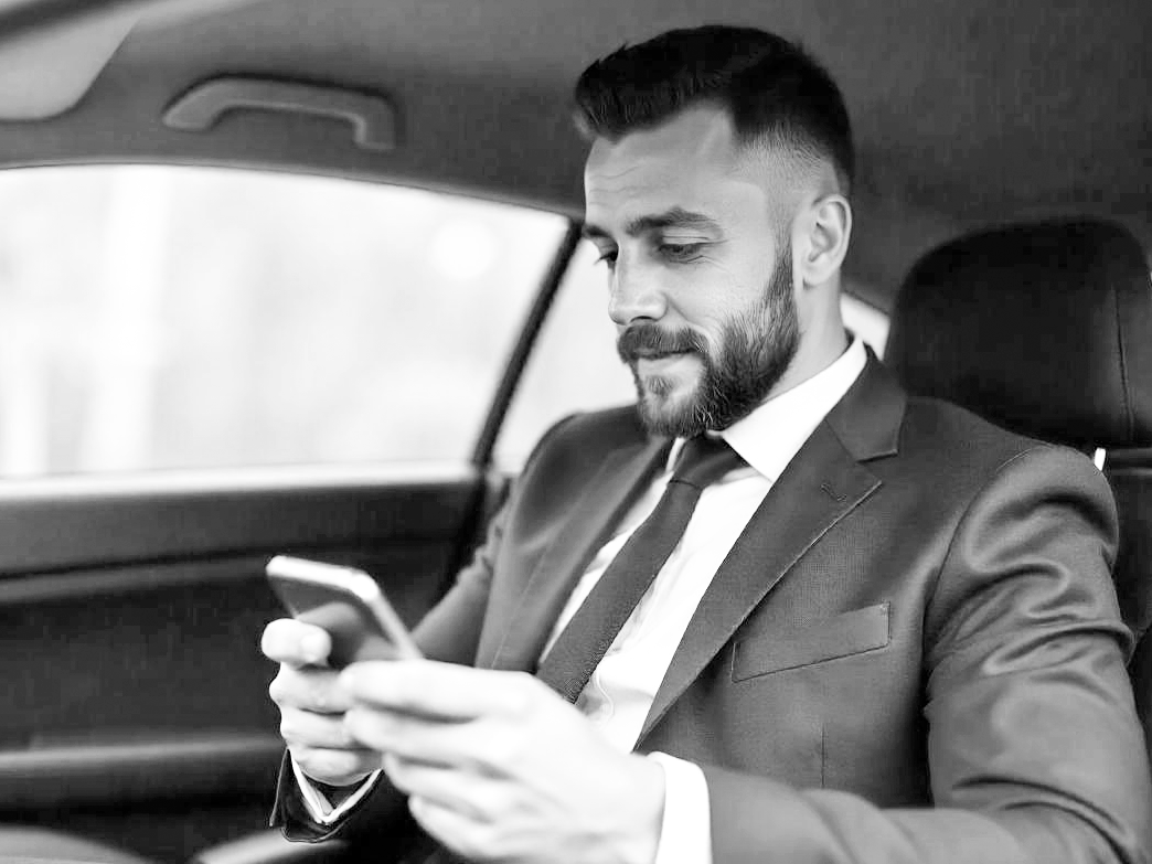 Homme en costume assis dans une voiture, regardant son téléphone.