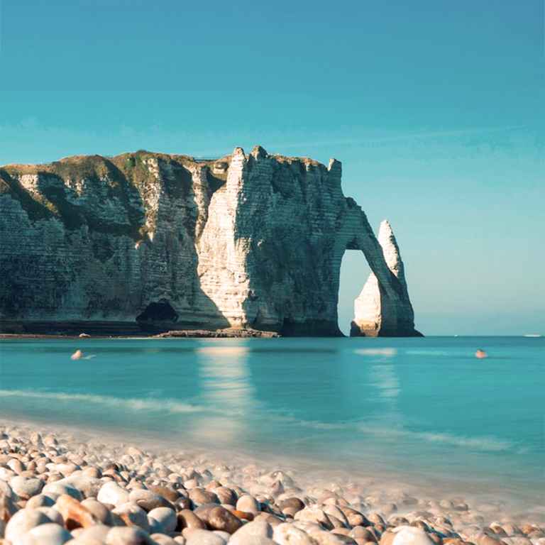 Côte avec une arche rocheuse surplombant une mer calme et un rivage de galets.
